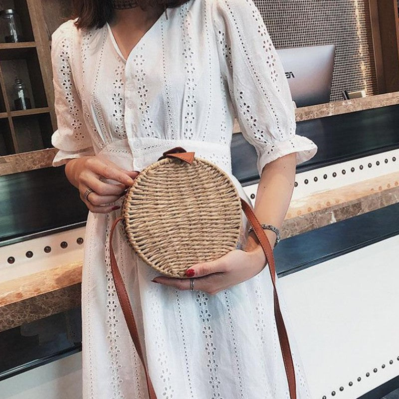 Person wearing a polka dot dress standing holding a round woven bag in her hands, in a sunlit outdoor area.