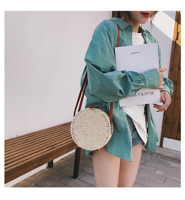 Person wearing a teal shirt with a round woven bag over their shoulder, standing in a sunlit outdoor area.