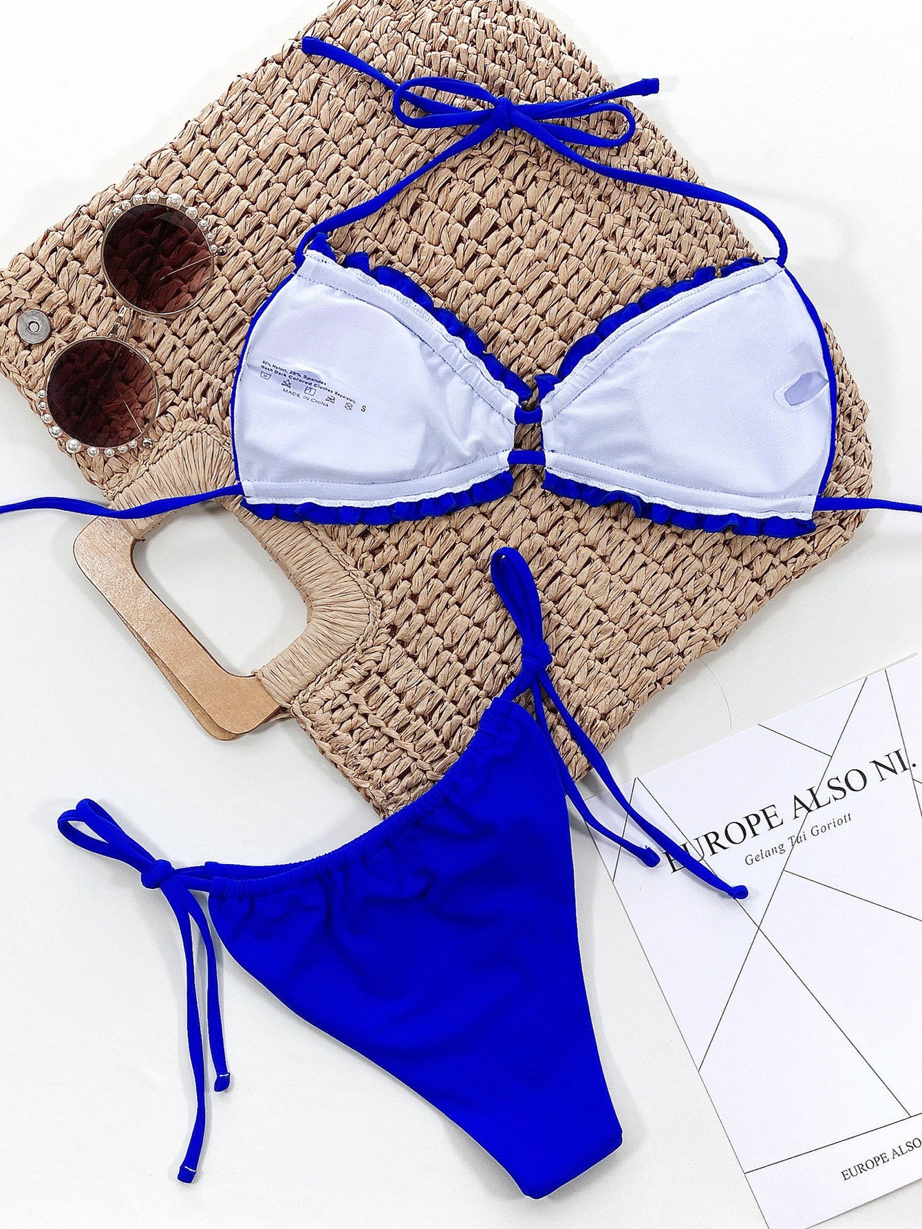 White bikini top with blue trim and matching bottom on a white surface with sunglasses and paper.