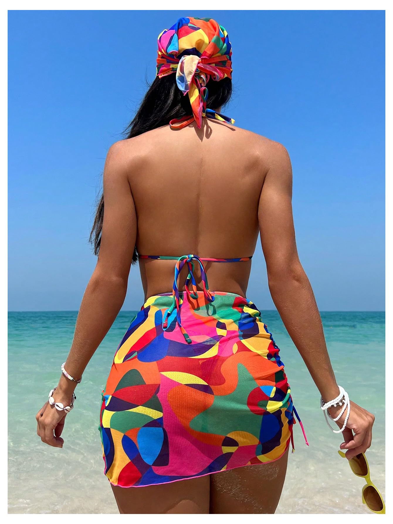 Woman in a colorful outfit standing on a beach with clear blue water and sky.