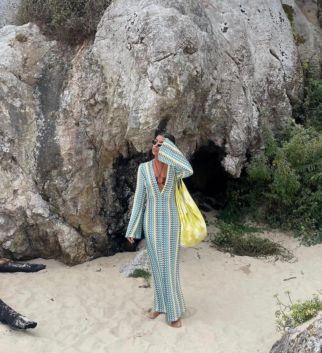 Person in a patterned dress standing in front of large rocks on a beach.