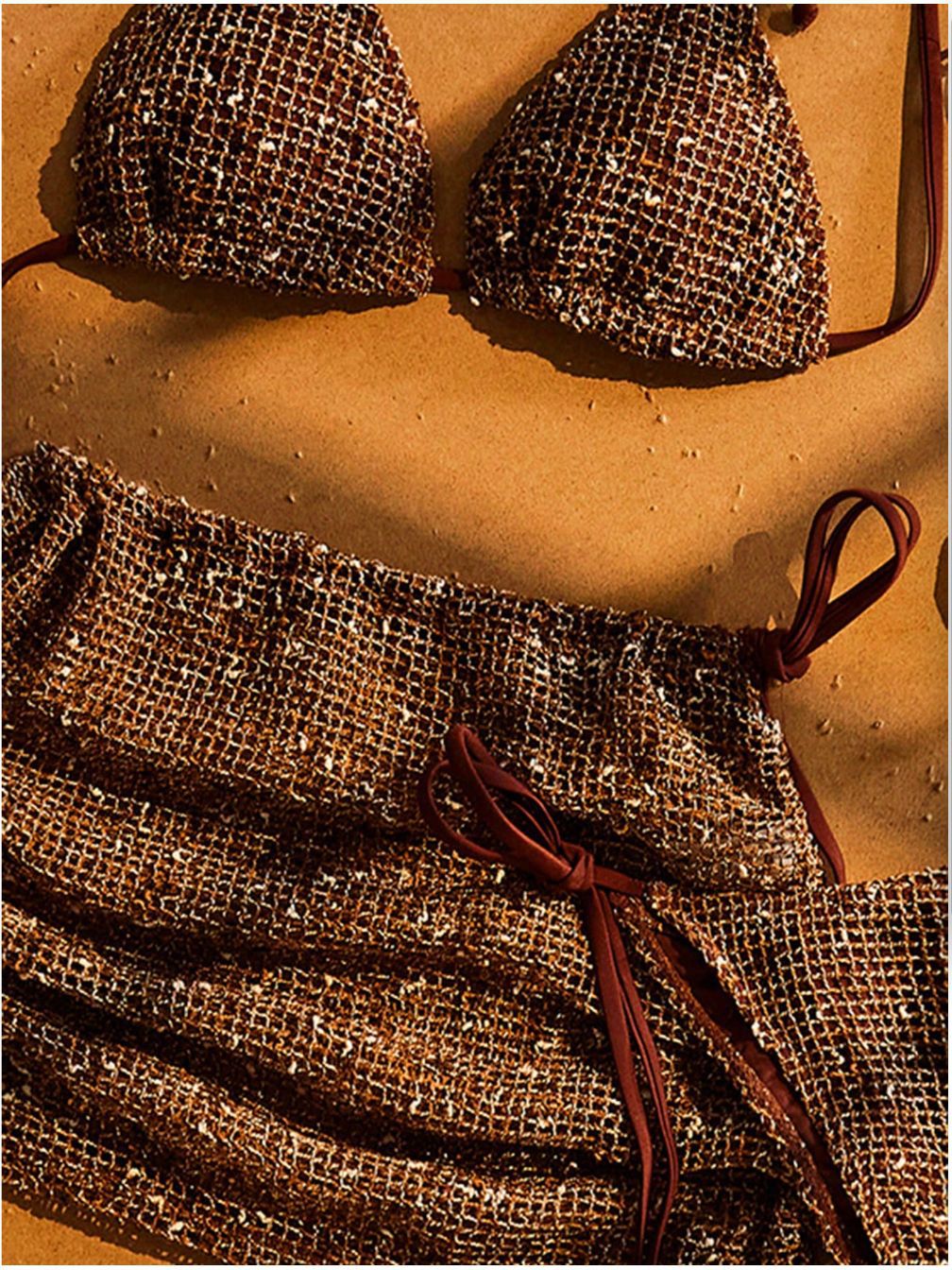 A close-up of a metallic bronze bikini set with a textured, woven mesh-like pattern and dark brown ties, laid flat on a sandy beige background.