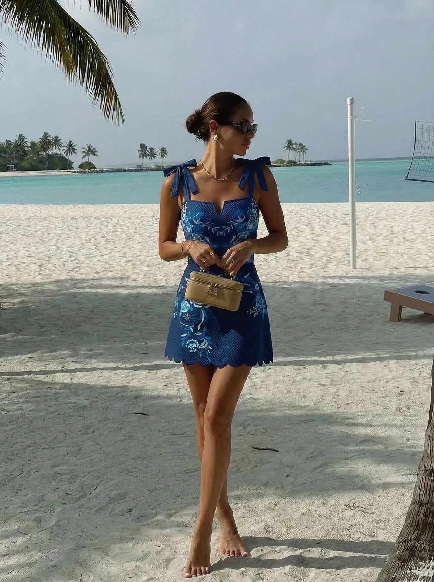 A woman stands on a sandy beach wearing a blue sleeveless dress with a white floral print, holding a small tan handbag.