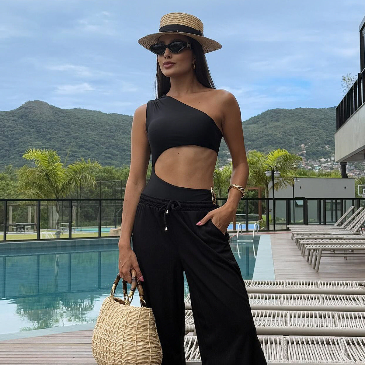 Woman in black one-shoulder top and wide-leg pants with a straw hat and bag by a pool with mountains in the background. Sold at PeachyBeanz.