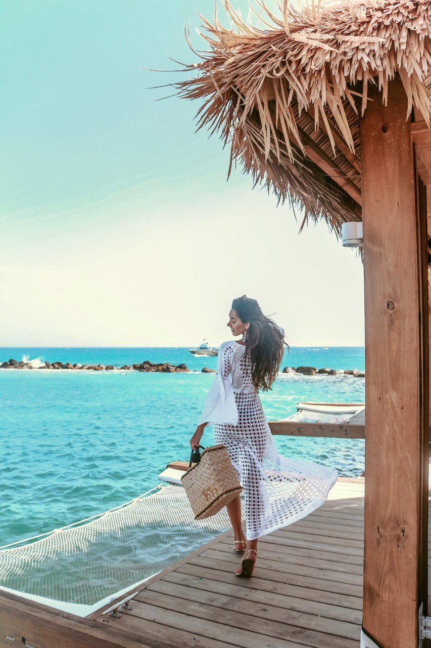 Woman walking on a wooden deck by the ocean with a thatched roof structure. Sold at PeachyBeanz.