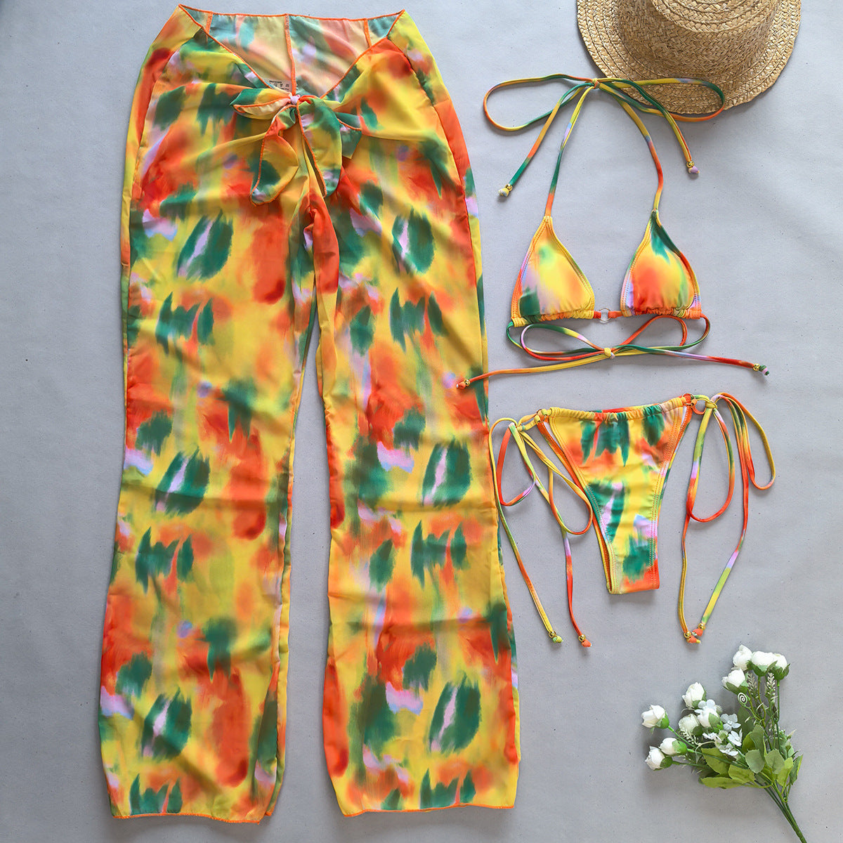 Colorful tie-dye pants and bikini set on a gray surface with a straw hat and flowers.