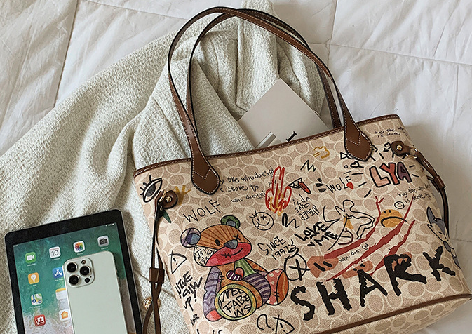A beige tote bag with brown leather handles and a cartoon bear illustration, styled on a white textured blanket next to a tablet.