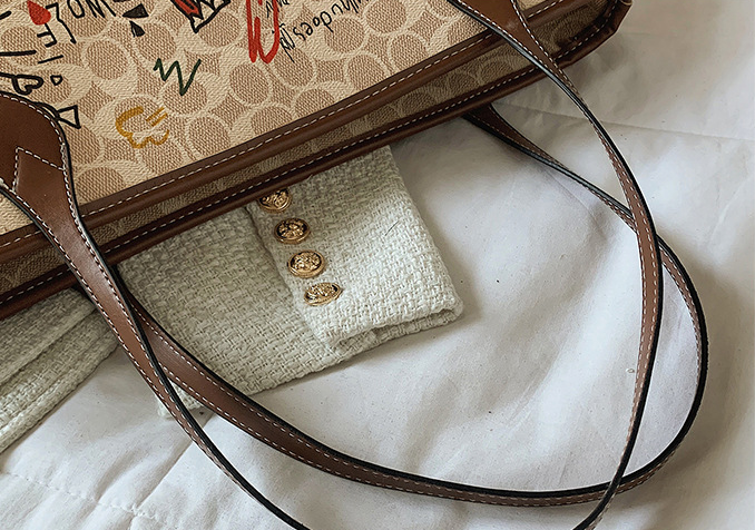A close-up of a beige and brown patterned tote bag with dark brown leather straps, laid on a white textured fabric surface next to a folded cream tweed garment with gold buttons.