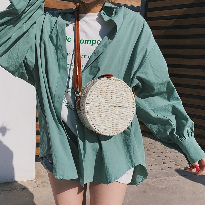 Person wearing a teal shirt with a round woven bag over their shoulder, standing in a sunlit outdoor area.