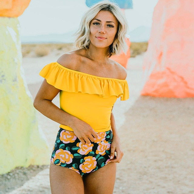 Woman wearing a yellow top and blue floral shorts standing against a rocky background