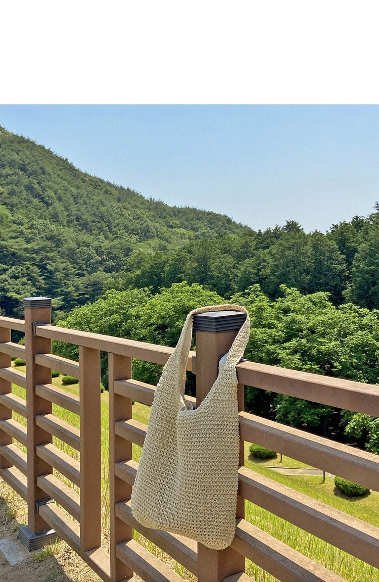 Beige woven bag hanging on a wooden railing with a scenic background of green hills and trees.