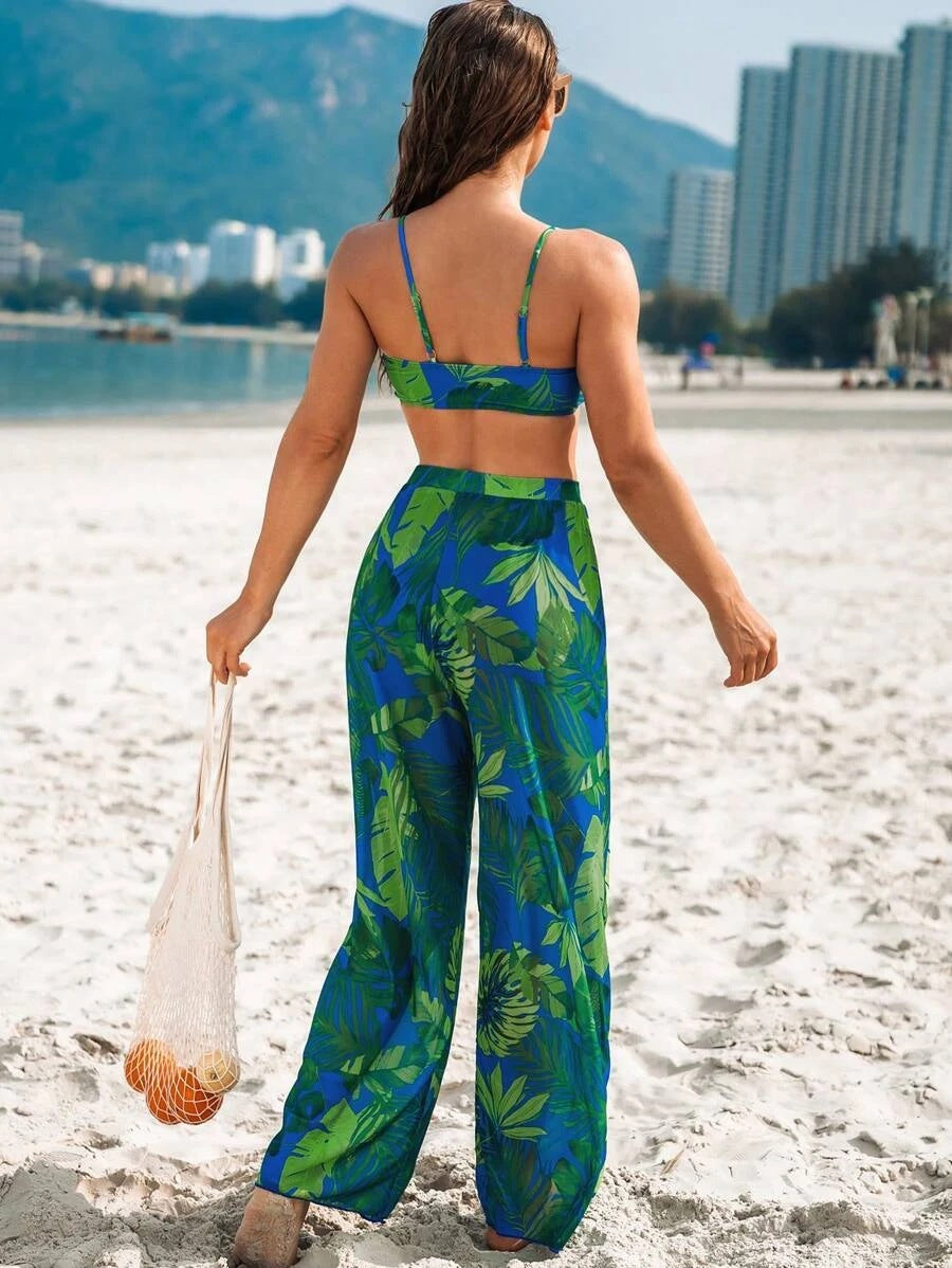 A woman stands on a sandy beach facing away from the camera, wearing a matching two-piece swimsuit with a blue and green tropical leaf print. She is holding a white mesh bag with a small orange fish.