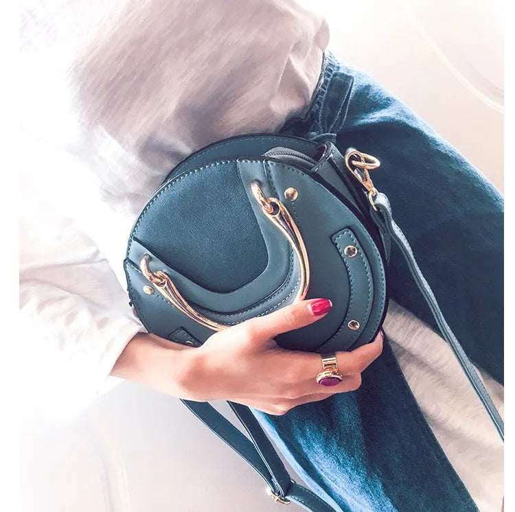Hand holding a teal handbag with gold accents, wearing a white shirt and blue jeans.