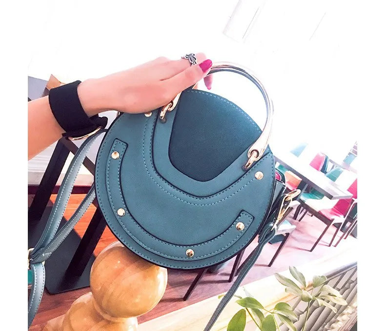Teal handbag held by a hand with pink nail polish, blurred background. Sold at PeachyBeanz.