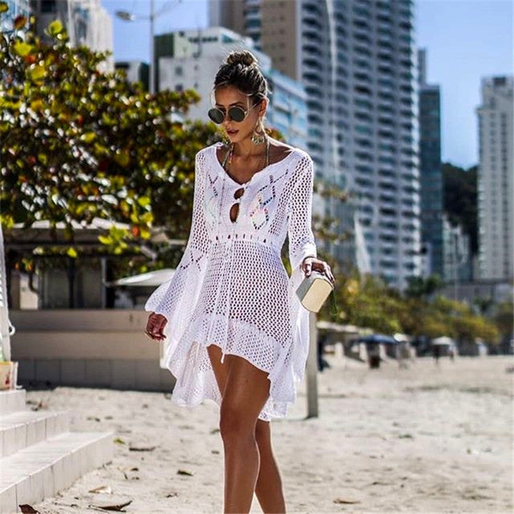 Woman in a white crochet dress standing on a beach with city buildings in the background