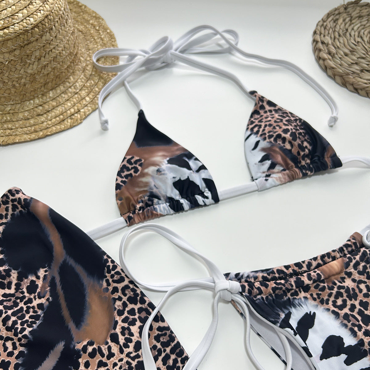 Leopard print bikini set with white straps on a white surface, accompanied by a straw hat and sunglasses.
