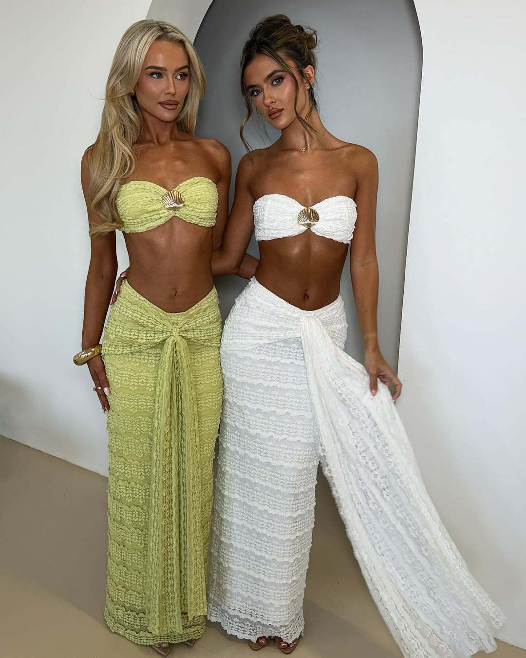 Two women wearing strapless tops and long skirts against a plain background