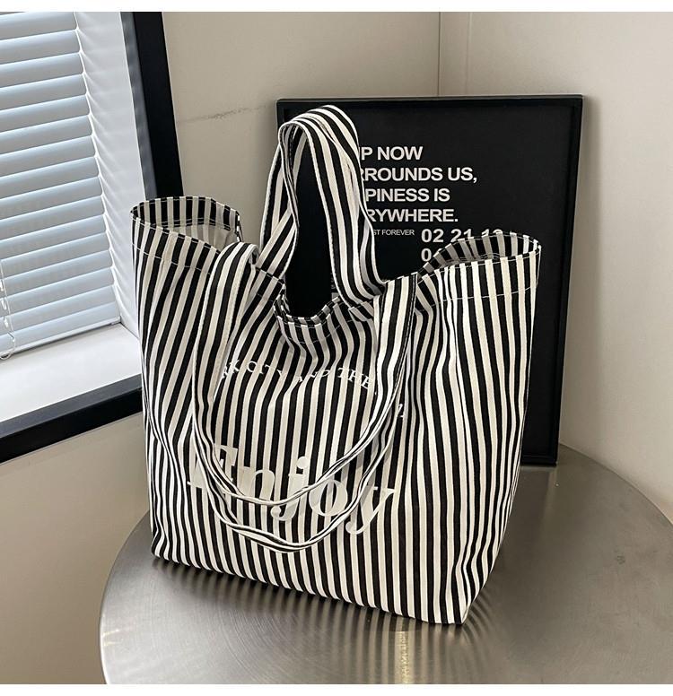 A black and white vertically striped canvas tote bag with white text reading 'The Boy' on the front, displayed on a metallic table next to a window with blinds.