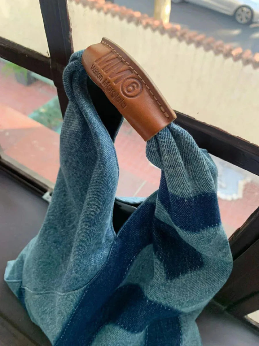 A close-up of a woven tote bag with a blue and navy chevron pattern and a brown leather handle, hanging near a window.